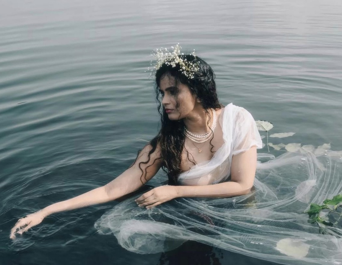 A woman with a baby's-breath flower crown and pearl necklace sits chest-deep in rippling grey-green water, sheer white fabric floating around her as she trails one arm across the surface.