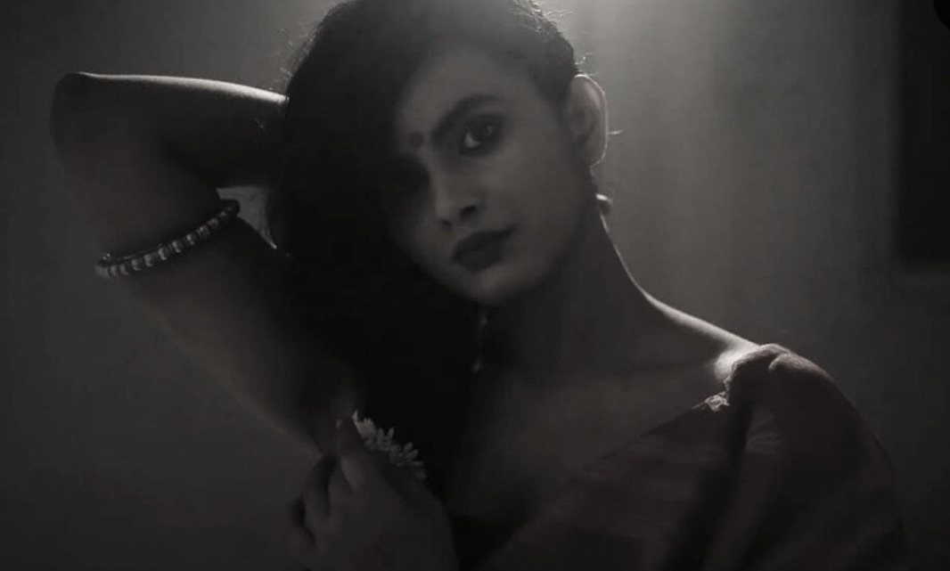 A dimly lit portrait of a woman with a bindi and dark lipstick raising one arm behind her head, a beaded bracelet on her wrist, in a shaft of smoky light.