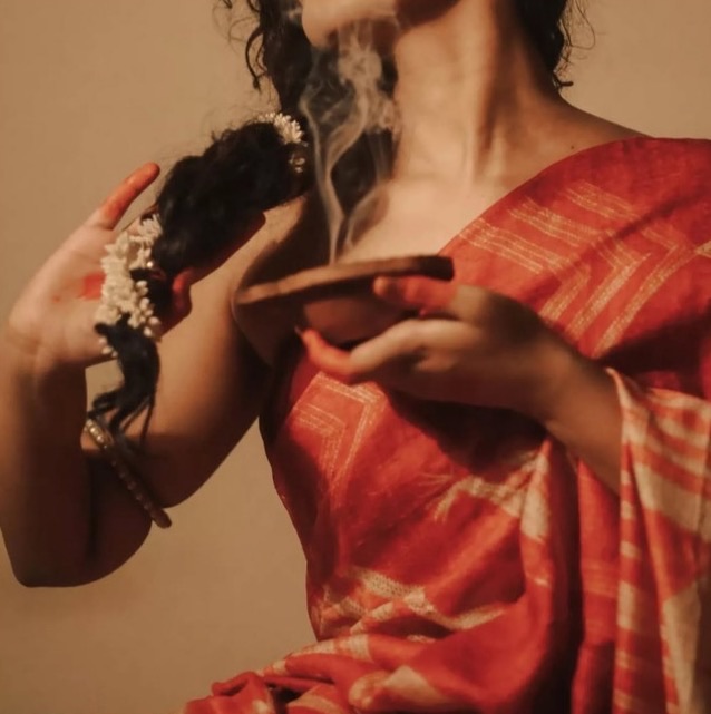 A woman in a red-and-cream patterned sari holds a smoking clay vessel below her chin while lifting her jasmine-wrapped braid, smoke curling up past her face.