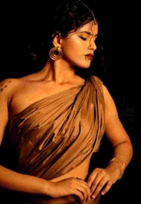 A standing woman wrapped in pleated bronze drapery, eyes closed in profile with a spiral gold earring, lit warmly against a black background.