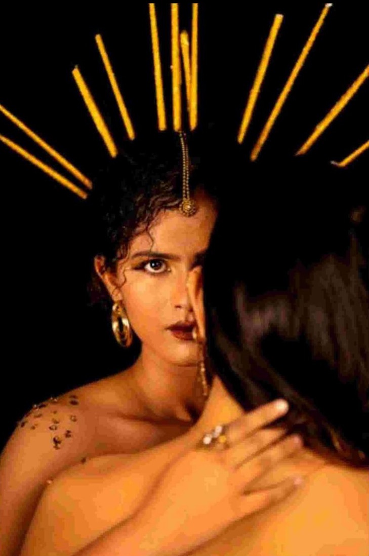 Styled photograph of a woman in a bronze drape before a crown of golden rays on black