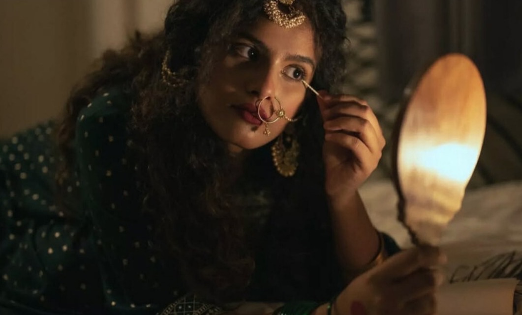 In candlelight, a woman wearing a dark green polka-dot kurta, gold nose ring and headpiece touches her eye makeup while holding a glowing wooden hand mirror.