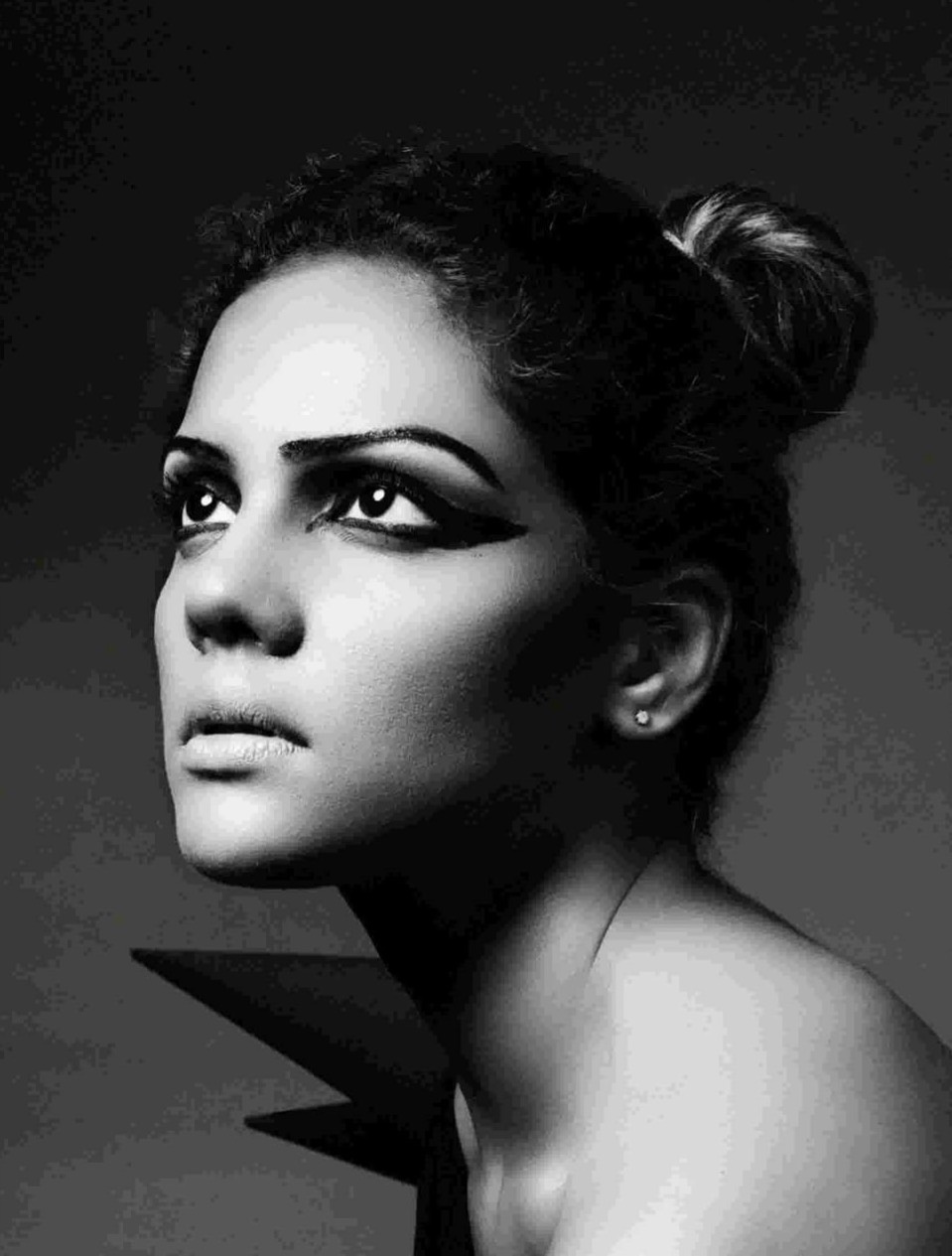 A black-and-white profile portrait of a woman with her hair in a bun, sharp graphic black eye makeup, and a black collar of conical spikes rising at her shoulder.