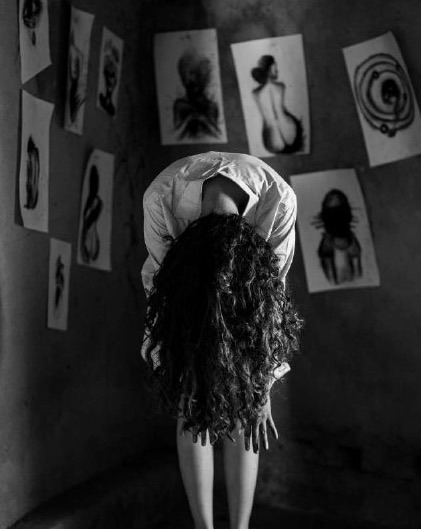 The artist bends forward from the waist with her curly hair spilling over her face, standing in a corner room hung with her charcoal figure drawings.