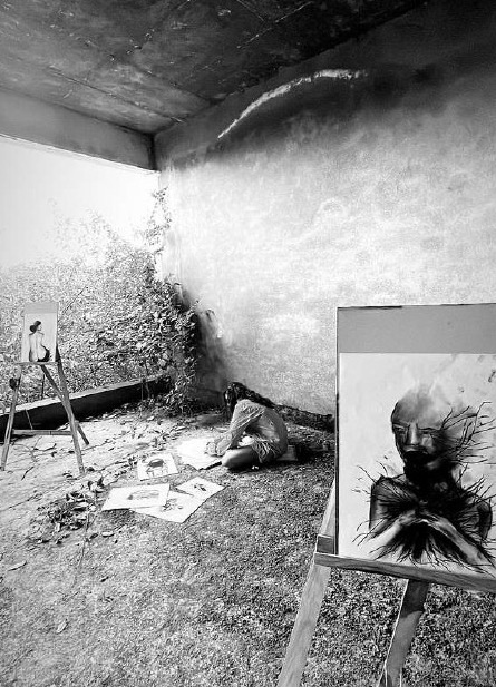 The artist sits on overgrown grass in a derelict open-walled room, working on drawings spread around her, flanked by two easels holding charcoal portraits.