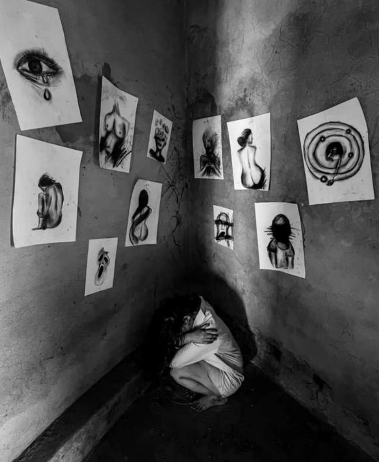 The artist crouches hugging her knees in the dark corner of a room whose two converging walls are pinned with charcoal drawings of a weeping eye, torsos and distorted figures.