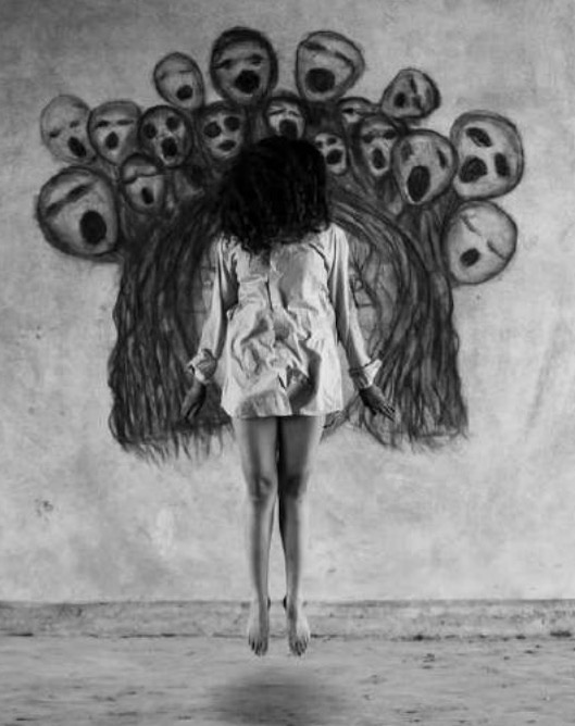 The artist floats barefoot in mid-air, seen from behind, in front of the charcoal wall mural of screaming faces, her real hair merging with the mural's drawn hair.